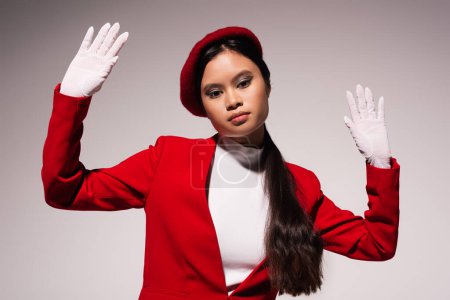 Trendy asian model in jacket and white gloves looking at camera isolated on grey 