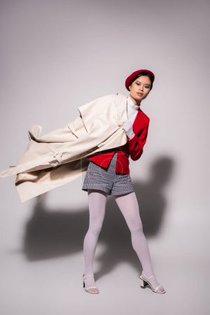 Trendy asian woman in beret wearing trench coat on grey background 