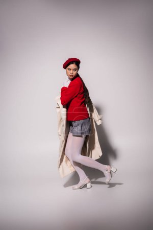 Full length of stylish asian woman in red jacket and beret holding trench coat on grey background 