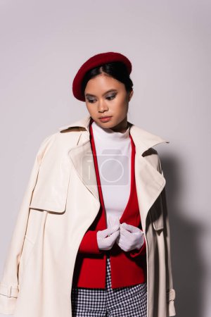 Portrait of fashionable asian woman in beret and trench coat standing on grey background 