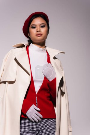 Low angle view of stylish asian woman in red beret and beige trench coat looking at camera isolated on grey 