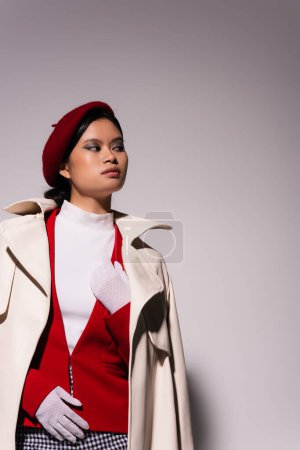 Pretty young asian woman in red jacket and trench coat looking away on grey background 