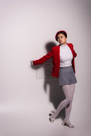 Trendy asian woman in checkered shorts and red jacket standing on grey background 