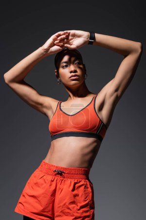 Foto de Young african american sportswoman in red sports bra posing with raised hands isolated on grey - Imagen libre de derechos