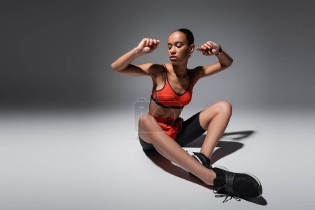 Téléchargez les photos : Full length of brunette african american woman sitting in shorts and sports bra sitting on grey background - en image libre de droit