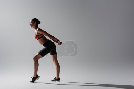 Photo for Full length of tattooed african american woman in sports bra and shorts stretching on grey - Royalty Free Image