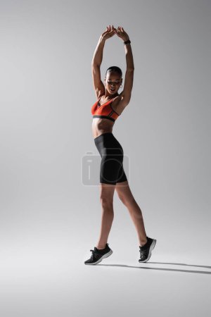 Photo for Full length of tattooed african american woman in sports bra and shorts stretching with raised hands on grey - Royalty Free Image