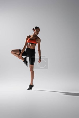 Foto de Full length of brunette african american woman in sports bra and shorts stretching leg on grey - Imagen libre de derechos