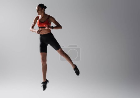 full length of tattooed african american woman in sports bra and shorts jumping on grey background 