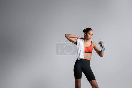 Foto de Young african american woman in sports bra and bike shorts standing with sports bottle and towel on grey background - Imagen libre de derechos