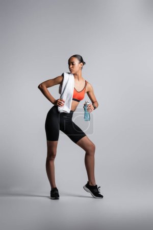full length of african american woman in sports bra and bike shorts standing with sports bottle and towel on grey background 