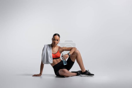 young african american woman in sports bra and bike shorts sitting with sports bottle and towel on grey background 