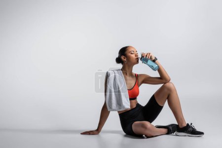 Foto de Young african american woman in sports bra and bike shorts sitting with towel and drinking water on grey background - Imagen libre de derechos