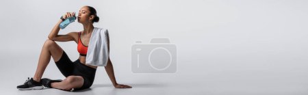 Photo for Young african american woman in sports bra and bike shorts sitting with towel and drinking water on grey, banner - Royalty Free Image