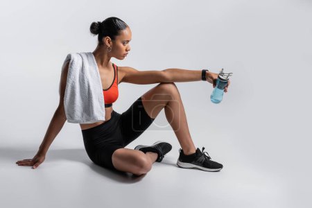 Téléchargez les photos : Side view of young african american woman in sports bra and bike shorts sitting with sports bottle and towel on grey background - en image libre de droit