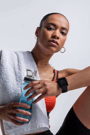 Téléchargez les photos : Young african american woman in sports bra sitting with sports bottle and towel isolated on grey - en image libre de droit