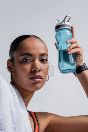 Téléchargez les photos : Brunette african american woman in sports bra holding sports bottle with water isolated on grey - en image libre de droit