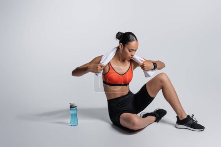Foto de Full length of young african american sportswoman sitting with sports bottle and towel on grey background - Imagen libre de derechos