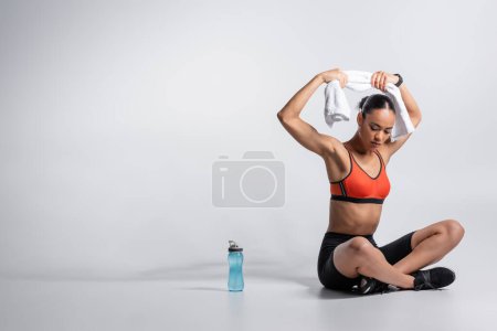 full length of young african american sportswoman sitting and holding towel near sports bottle on grey background 