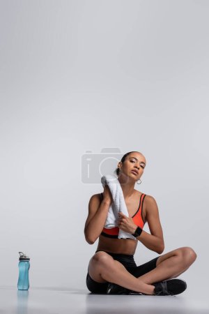 full length of brunette african american sportswoman sitting with towel near sports bottle on grey 