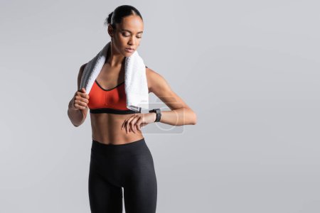 Foto de Brunette african american sportswoman in red sports bra standing with towel and looking at fitness tracker isolated on grey - Imagen libre de derechos
