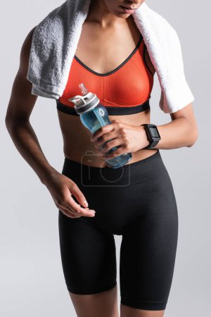Foto de Cropped view of young african american sportswoman standing with towel and sports bottle isolated on grey - Imagen libre de derechos