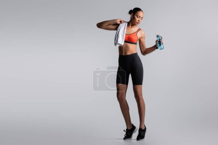 full length of young african american sportswoman standing with towel near sports bottle on grey background 