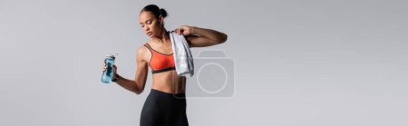 Foto de Young african american sportswoman standing with towel near sports bottle isolated on grey, banner - Imagen libre de derechos