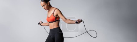 sportive african american woman exercising with skipping rope on grey background, banner 