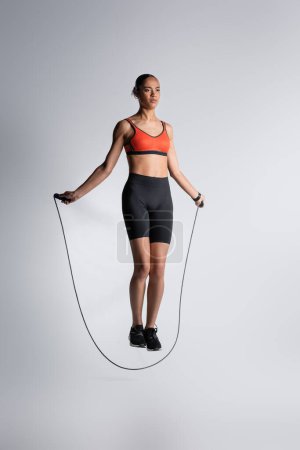 Photo for Full length of african american sportswoman exercising with skipping rope on grey - Royalty Free Image
