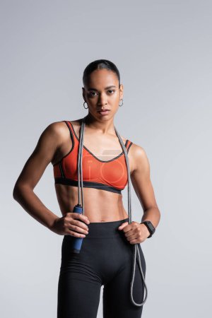 Foto de Young african american sportswoman standing with skipping rope and looking at camera isolated on grey - Imagen libre de derechos