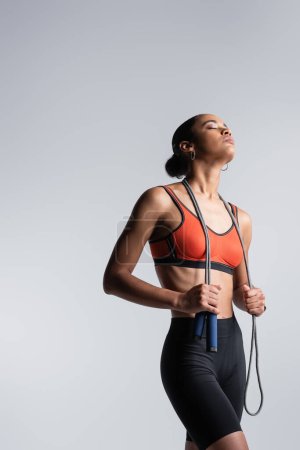 Foto de Young african american sportswoman with closed eyes holding skipping rope isolated on grey - Imagen libre de derechos