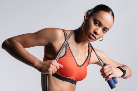 Photo for Brunette african american sportswoman in red sports bra holding skipping rope isolated on grey - Royalty Free Image