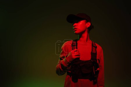 young african american woman in baseball cap and sweatshirt standing on green background with red light 