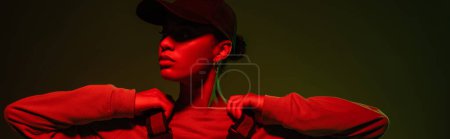 african american woman in sweatshirt and baseball cap posing on green with red light, banner 