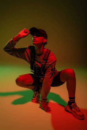 full length of african american woman in sweatshirt and vest sitting while adjusting baseball cap on green with red light 