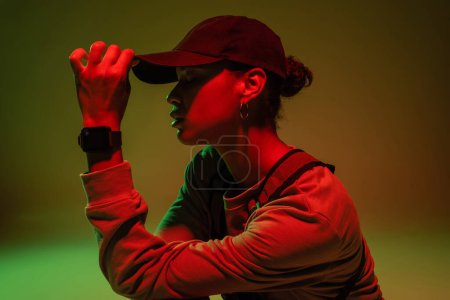 Photo for Side view of african american woman in sweatshirt adjusting baseball cap on green with red light - Royalty Free Image