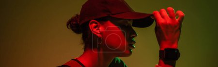 Photo for Side view of african american woman in sweatshirt adjusting baseball cap on green with red light, banner - Royalty Free Image