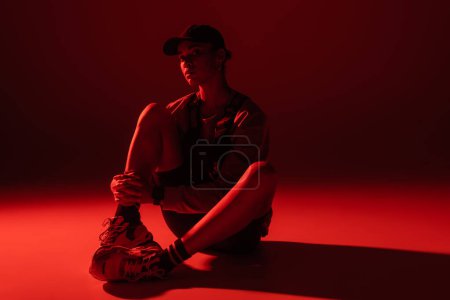 full length of young african american woman in baseball cap and sweatshirt sitting on dark red background 