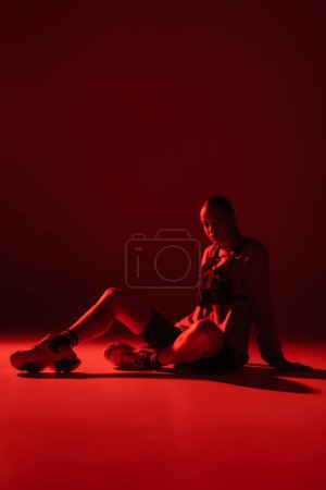 full length of young african american woman in bike shorts and sweatshirt sitting on dark red background 