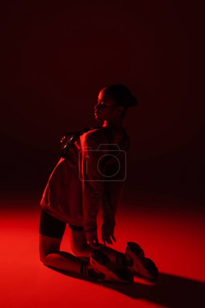 full length of young african american woman in bike shorts and sweatshirt posing on dark red background 