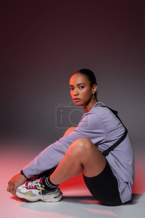 full length of young african american woman in bike shorts and sweatshirt sitting on grey with red light 
