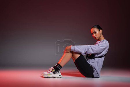 Photo for Full length of african american woman in bike shorts and sweatshirt sitting on grey background with red light - Royalty Free Image