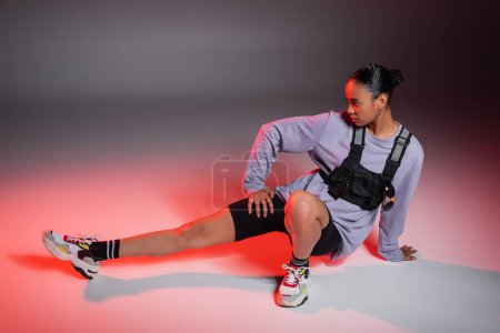 full length of african american woman in bike shorts and sweatshirt posing on grey background with red light 