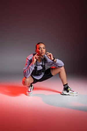 Photo pour Full length of african american woman in bike shorts and sweatshirt sitting while wearing sunglasses on grey background with red light - image libre de droit