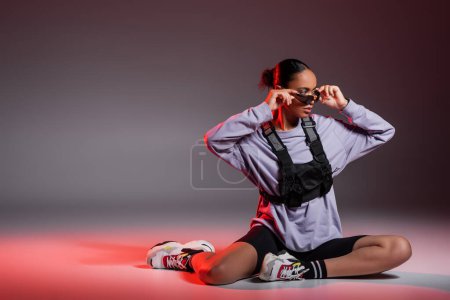 full length of african american woman in bike shorts and sweatshirt wearing sunglasses on grey background with red light 