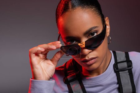 portrait of african american woman in sweatshirt and stylish sunglasses looking at camera on grey with red light 