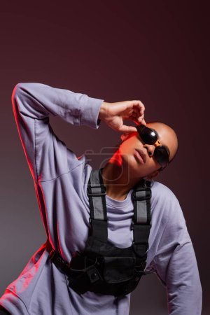 Photo for Portrait of african american woman in sweatshirt adjusting trendy sunglasses on grey with red light - Royalty Free Image
