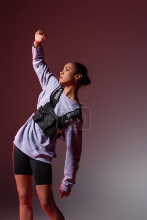 Photo for Young african american woman in sweatshirt and bike shorts posing with raised hand on grey and purple - Royalty Free Image