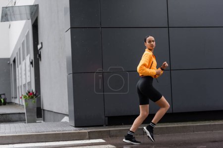 Foto de Full length of brunette african american sportswoman in bike shorts and yellow puffer jacket running outside - Imagen libre de derechos
